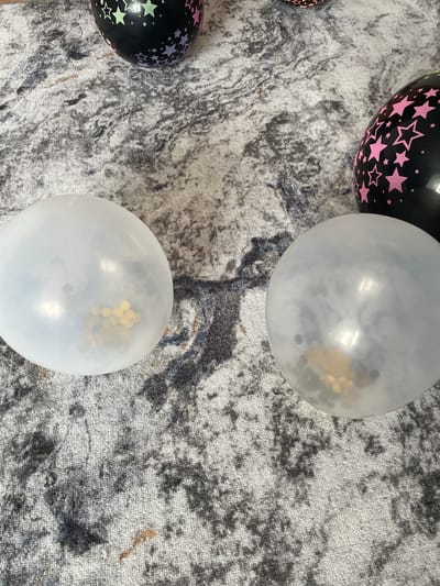 The image shows several balloons scattered on a carpeted surface. In the foreground, there are two clear balloons, each containing some confetti. One balloon is on the left, and the other is on the right. In the background, there are two black balloons with star patterns. One has green and purple stars, and the other has pink stars. The carpet has a marbled pattern of gray, black, and white. The scene appears to be indoors, possibly in a home or event venue. There are no people or any specific landmarks visible that would allow for determination of a precise location in Азершчынскі сельскі Савет, Belarus.