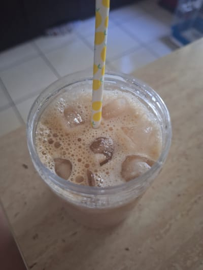The image shows a close-up, top-down view of an iced beverage in a clear plastic cup. The drink is a light brown color, topped with a frothy layer of small bubbles and several ice cubes. A yellow and white striped straw with a lemon pattern is inserted into the drink. The cup is placed on a light brown, speckled surface that appears to be a table. In the blurred background, a tiled surface and a darker, out-of-focus area are visible. The lighting suggests it is daytime. There is no visible text in the image. The overall impression is of a refreshing drink ready to be consumed.