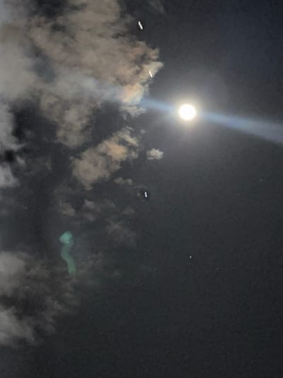 This image depicts a nighttime sky over Beit Yitshak Sha'ar Hefer, Israel. A prominent, bright, circular full moon is visible in the upper right quadrant, emitting a strong, horizontally extending light ray towards the left. Dark, broken cloud formations occupy the left side and partially obscure the upper and central portions of the frame. Some cloud edges exhibit iridescent coloration, including hues of yellow, orange, and faint spectral bands.
Multiple elongated, bright white, cylindrical or pill-shaped objects are distributed across the sky. Two such objects are positioned vertically in the upper left, with another located slightly below and to their right. A fourth similar object is present in the mid-right area, approximately level with the moon's horizontal light ray. A faint, small white dot is visible towards the lower right.
A distinct, luminous, greenish-blue, serpentine form is observed in the lower-left quadrant, partially integrated with the cloud structure. This form features a brighter, bulbous upper section tapering into a more attenuated, curved tail. No discernible ground features, architecture, or human figures are present.