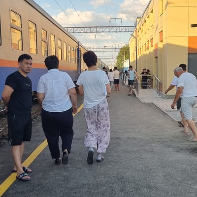 The image depicts a train station platform in the city of Arys, Kazakhstan. In the foreground, a man in a black t-shirt and shorts stands facing the camera, while two people, one in a white shirt and dark pants and another in a white t-shirt and floral-patterned pants, walk away from the camera. A train is visible on the left side of the image, and a yellow line marks the edge of the platform. In the background, more people are seen on the platform near a light-yellow building. Overhead, there are train tracks and electrical wires. The sky is partly cloudy.