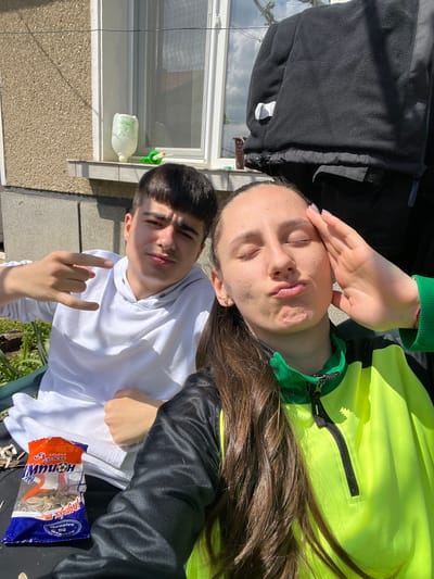 Outside a building in Hrishteni, Bulgaria, a teenager wearing a white hoodie makes a devil horn hand gesture next to another teenager wearing a bright yellow jacket and leaning back with eyes closed and one hand on their forehead. The teenagers are positioned next to a packet of sunflower seeds. Outside, the building has a window with a bottle attached below. A black jacket is partially visible at the top.