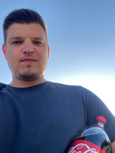The image shows a close-up selfie of a young man holding a bottle of Coca-Cola. He is wearing a dark blue t-shirt and has short brown hair and a light beard. The background is a clear blue sky, indicating an outdoor setting. The man is looking towards the camera, and the Coca-Cola bottle is positioned in his right arm, resting against his body. The lighting suggests it is daytime, possibly late afternoon due to the warm glow on his face and the sky. There is no identifiable information about the location in Yablunivka, Ukraine within the image itself.