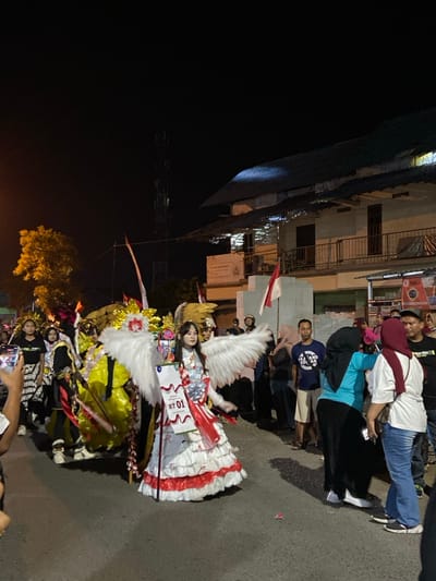 The image captures a nighttime procession occurring on an asphalt street in Kedungbanteng, Indonesia. A central figure, appearing to be a young female, is dressed in an elaborate white and red costume featuring large white feathered wings, a multi-layered ruffled dress, and a white sash emblazoned with "RT 01" and Indonesian flag designs. She holds a staff or pole in her left hand and faces slightly right. Behind her, other parade participants in complex, colorful costumes (predominantly yellow, black, and red) are visible. On the right side of the street, a crowd of spectators, dressed in casual clothing including hijabs, observes the procession. An Indonesian flag is displayed on a pole affixed to a white, brick-like structure among the spectators. In the background, a two-story building with a dark, sloping roof, balconies, and visible signage stands on the right. A tall telecommunications tower is silhouetted against the dark sky behind the building. On the left, an artificially lit tree with yellow foliage is present. The scene is illuminated by various artificial light sources. A hand holding a mobile phone, directed towards the main subject, is visible in the lower left foreground.