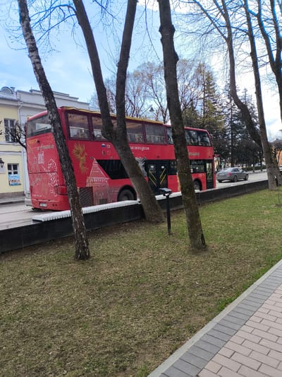 A vibrant red double-decker tour bus is the central subject of this daytime image, seen from its rear-left side. It is positioned on a paved road, partially obscured by several bare-branched trees in the foreground. The bare trees and muted, patchy grass suggest a late winter or early spring setting under a bright, possibly overcast, sky.
The bus is decorated with white line art illustrations and Cyrillic text. On its rear, the words "ЭКСКУРСИЯ по Смоленску" (Excursion in Smolensk) are clearly visible, indicating its purpose as a sightseeing tour in the Russian city of Smolensk. Other details include a QR code, a cartoon character with luggage, a stylized depiction of a historical building (resembling a fortress or kremlin tower), and a golden phoenix or dragon motif on the bus side. Small Russian and British flags are displayed near the upper rear windows. The visible license plate, A 902 AX 67, includes the region code "67" which corresponds to Smolensk Oblast, further confirming the location context.
To the right of the bus, a grey sedan is driving on the road. The foreground features a strip of dry, patchy grass leading to a neat paved path constructed with light and dark grey interlocking bricks. Behind the bus, traditional-style buildings with light-colored facades and arched windows line the street. A sign on one of these buildings partially reads "Экскурсии на теплоходе" (Boat excursions). The scene captures an urban environment, likely in Smolensk, Russia, during a calm daytime period.