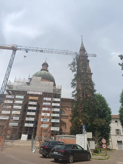 The image shows the Duomo of Casalmaggiore, Italy, under renovation. A large construction crane looms over the dome, which is covered in scaffolding and white sheeting. Numerous banners, advertising local businesses and organizations, are attached to the scaffolding. The bell tower of the Duomo, with a large tree growing up its side, is visible to the right of the dome. In the foreground, two black cars are parked on a street in front of the Duomo. The sky is overcast.