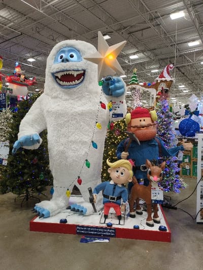 A large holiday display features iconic characters. A smiling Abominable Snowman towers, illuminated by a star on its head and string lights. Above him are animated figures of Yukon Cornelius, Rudolph, and Hermey the Elf, all on a snow-covered base. The display is surrounded by decorated Christmas trees and holiday decorations, suggesting it's in a retail environment. A sign advises against climbing on the display.