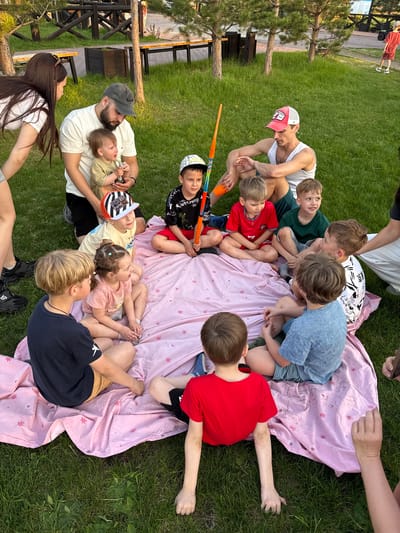 A group of people are gathered on a grassy lawn in what appears to be an outdoor setting. The main group consists of adults and children sitting around a pink blanket with flower designs. Children of varying ages are clustered together, some are sitting on the blanket while others are sitting around the perimeter. An adult, possibly a father figure, is holding a baby while conversing with a woman who stands nearby. A wooden picnic table is visible in the background, along with the trees and a body of water. The location is Кузнецово, Russia.