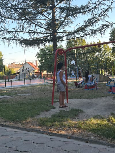 This media file depicts a scene at a playground in Kamianka-Buzka, Ukraine. The primary subjects are people, including at least two children on swings and one adult near a swing set. The setting is an outdoor playground with a red swing set in the foreground. Behind the swing set, there is a fenced-off area that appears to be a sports court, possibly for basketball, with a backboard visible. Further in the background, there are buildings with red roofs and trees.
The main activity is children playing on the swings. The adult in the foreground seems to be interacting with or observing the children. The time of day appears to be late afternoon or early evening, as indicated by the warm lighting and long shadows cast on the ground. The weather seems clear and sunny.
Notable details include the textured pavement on the right side of the image and the sandy surface of the playground. The trees in the foreground are dense and leafy. There is no visible text in the media file. The overall mood is relaxed and casual, typical of a playground setting.
