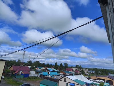 This is a high-angle, medium-shot, eye-level, outdoor, daytime, overcast photograph of a rural village in Milkovo, Russia. The sky is bright blue with scattered, puffy white clouds. Black power lines stretch diagonally across the frame from the upper right to the lower left. In the foreground, the edge of a white building with vertical siding is visible on the right. Below the power lines, the scene depicts a collection of residential houses and outbuildings with a variety of roof colors, including red, blue, green, and brown. Trees line the background. Two tall, lattice-style communication towers are visible in the middle ground to the left of center. Fences, made of various materials like wood and metal, delineate property lines. Two vehicles, a white car and a dark-colored sedan, are parked. The overall impression is that of a typical, somewhat rustic settlement.