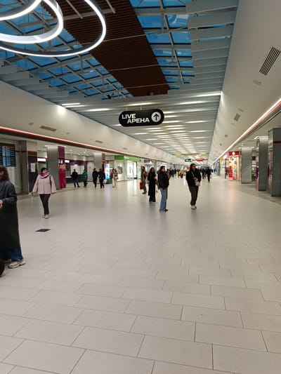 The image depicts an indoor pedestrian walkway, likely within a shopping mall or transportation hub, situated in Novoivanovskoe, Russia. The floor is covered with light-colored rectangular tiles, arranged in a running bond pattern. Overhead, a modern ceiling structure features linear lighting panels and a section with a blue-tinted glass roof, partially obscured by a dark brown, rectangular panel. Circular lighting fixtures are also visible.

People of varying ages and attires are walking along the corridor in both directions. Shops line both sides of the walkway, with retail displays and signage visible. A black sign with white text "LIVE АРЕНА" and an upward arrow is suspended from the ceiling, serving as a directional marker. Red LED strips run along the upper part of the walls on both sides. Overall, the scene portrays a busy, modern, and well-lit interior space.