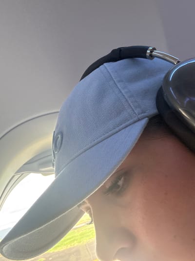 A person is seen from a close-up, skewed angle, wearing a light-colored baseball cap and black headphones. The individual appears to be on an airplane, with a window visible in the lower-left corner. Sunlight streams in, illuminating the scene and creating a hazy effect around the person's face. Focus is drawn to one eye and the bill of the cap.