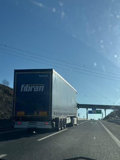 A semi-trailer truck is observed from a following vehicle on a multi-lane highway under a clear, bright blue sky. The truck's dark blue trailer features "fibran" in white lettering prominently on its rear and "fibran.pt" near the top right. Its rear bumper has "Krone" embossed. The road surface is asphalt, delineated by white lane markings. Hillsides or embankments line the highway on both the left and right. In the distance, an overhead gantry sign spans the highway, displaying a green sign with "IC5" and partially visible "MIRANDELA," indicating the location is on the IC5 motorway near Macedo de Cavaleiros, Portugal, heading towards Mirandela. Overhead power lines are visible across the sky. No individuals are present in the scene. The image exhibits characteristics consistent with being taken through a vehicle's windshield, including minor dust or spots.