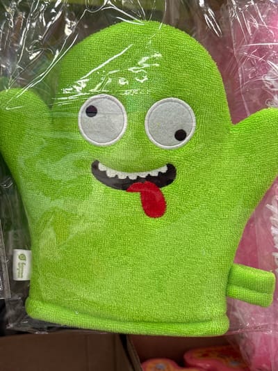 The image shows a close-up of a bright green, terrycloth bath mitt shaped like a cartoon monster. The mitt has two large, white circular eyes with black pupils. Its mouth is open, revealing jagged, black and white teeth and a red tongue sticking out. A small green loop or tab extends from the top of the mitt, likely for hanging. The mitt is enclosed in clear plastic packaging, which has some creases and reflections.
In the bottom right corner of the mitt, a small white tag is visible with green text. The text is in Cyrillic script and reads "Банные штучки" which translates to "Bath little things" or "Bath trinkets".
The background is blurred, but hints of other items are visible. To the left, a pink, possibly fabric or plastic item with a ruffled edge is partially seen, also covered in plastic. To the right, the edge of a cardboard box is visible, and behind it, a blurry pink object with a yellow and black pattern, possibly a toy or another product, can be discerned. The overall lighting is bright, suggesting an indoor setting, possibly a retail environment or storage area. No people are visible in the image. The mood conveyed by the bath mitt's design is playful and whimsical.