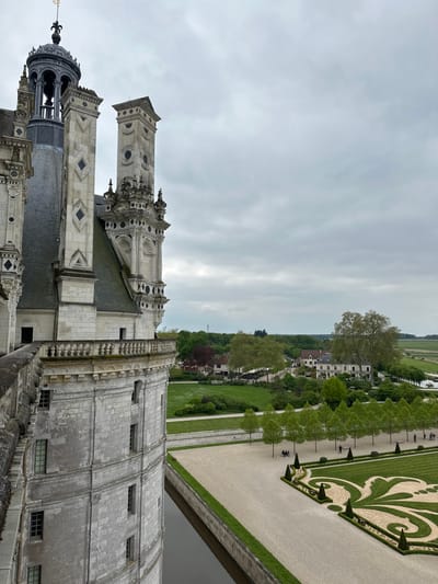 The image depicts a view from the upper level of the Château de Chambord, a Renaissance-era castle in Chambord, France (latitude: 47.616, longitude: 1.518). A portion of the castle's exterior is visible on the left, with a white stone facade adorned with windows, and a curved, ornate parapet. The castle's moat is directly below, with a narrow, rectangular channel of water visible in the foreground. Beyond the moat is a meticulously manicured landscape. A row of trees lines a pathway running parallel to the moat. A large, open space in the foreground is covered in a layer of gravel. This area features a complex design formed by a series of intersecting lines, arranged in a pattern that resembles a fleur-de-lis. There are a few individuals visible in the distance, some are seated on a stone bench while others are standing on the pathway. Beyond the manicured grounds, the countryside spreads out, with a large field to the right and a mix of trees and buildings in the background. The sky is overcast with gray clouds.