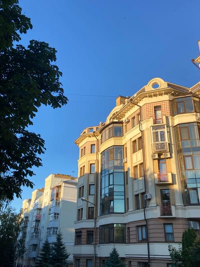 The image is an upward-facing shot of an urban street scene, dominated by a clear blue sky. In the foreground, dark green foliage from trees partially obscures the view of multi-story buildings. The buildings themselves are a mix of architectural styles, with cream-colored facades, brown paneling, and numerous windows, some of which are large and reflective, showing the sky. The sunlight casts long shadows, suggesting it is late afternoon or early morning.

One building, with a more ornate design, features a patterned facade with diamond-shaped tiles and decorative railings. Another, a lighter color, displays a series of balconies and many windows, some of which are covered by white blinds. A red sign is visible on one of the buildings. Small trees, possibly evergreens, are interspersed with the larger trees and buildings. Several small dark specks, possibly birds or insects, are visible in the sky.

The overall impression is one of a bright, clear day in a European city. The orientation of the shot, looking upwards, gives a sense of scale and grandeur to the architecture against the expansive sky.