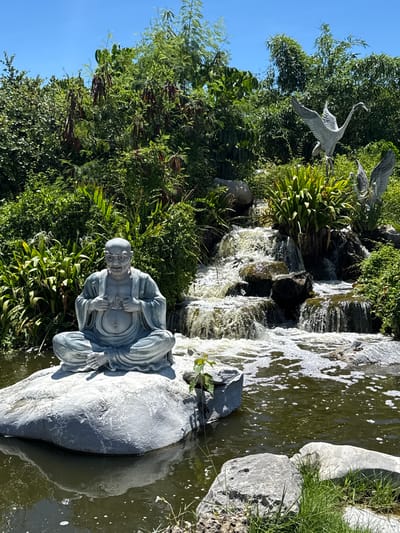 A statue of a smiling, bald, seated man in traditional garb rests on a large stone beside a pond and a waterfall in Bang Pu Subdistrict Municipality, Thailand. Two sculptures of cranes stand among lush green foliage and other natural elements. The sky is clear and blue.