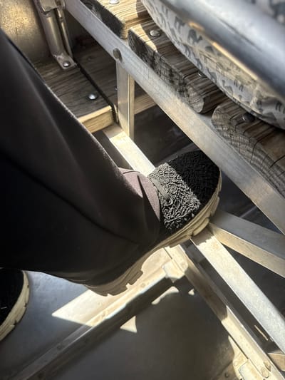 A close-up shot from a low angle depicts a person wearing black pants and shoes with a lace-like pattern on the top, standing on a ladder. The ladder appears to be made of metal and wood. The environment seems to be a building interior, possibly in Weston, United States. The lighting suggests natural light coming in from above.