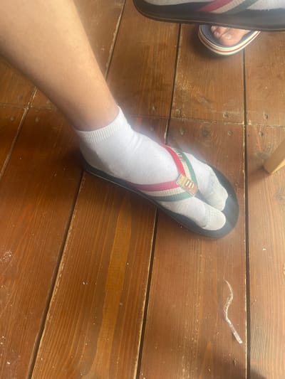The image shows a person's left foot, clad in a white sock and a black flip-flop with red, white, and green striped straps, resting on a wooden floor. The floorboards are narrow, stained dark brown, and show signs of wear, with visible nail heads and some dust. The foot appears to be positioned indoors, as indicated by the wooden flooring. There is no visual information within the image that can be used to determine its location in the city of Ryazantsevo, Russia.