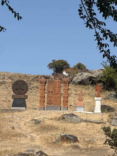 The image displays an outdoor scene in the city of Shirak, Armenia, featuring a collection of stone monuments on a dry, rocky hillside under a clear blue sky. In the foreground, from left to right, four distinct monument groupings are visible. The leftmost consists of a dark, intricately carved circular stone disk mounted on a dark rectangular pedestal with a gray base. Adjacent to it stands a three-part monument of red-brown stone, comprising a central vertical slab deeply carved with crosses and geometric patterns, flanked by two ornate pillar-like structures, all resting on a shared dark base. To the right, a smaller red-brown carved cross-stone (khachkar) is situated on a lighter rectangular base. The rightmost monument is a red-brown cross mounted on a tall, white rectangular pedestal. The ground is covered with sparse, dry, yellow-brown grass and scattered dark rocks; a faint dirt path is present in the lower-left quadrant. The background reveals a rising hill with similar dry vegetation, rocks, and several small green trees. Dark, leafy tree branches partially extend into the upper left and upper right corners of the frame. The scene is devoid of human presence or activity.