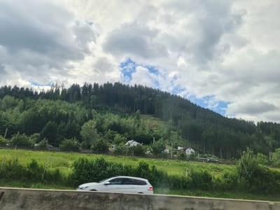 The image depicts a scenic view from a roadside, likely a highway, in Niklasdorf, Austria. The foreground features a concrete barrier. Beyond this, a grassy embankment slopes up towards a wooded hillside. A white station wagon is in motion on the right side of the road in the mid-ground, its side visible. A dark-colored vehicle is also visible further down the road. Scattered houses with white walls and dark roofs are nestled amongst the trees on the lower slopes of the hill. The hillside is densely covered with a mix of evergreen and deciduous trees, with some evidence of logging activity. Power lines and transmission towers are visible on the far left of the hill. The sky above is filled with dramatic, heavy grey clouds, interspersed with patches of blue sky. The overall impression is a natural landscape viewed from a transportation route.