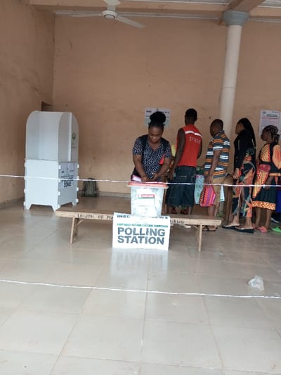 An indoor polling station in Ajalli, Nigeria, features light brown walls and a beige tiled floor. A white, three-bladed ceiling fan is mounted overhead. On the far left, two white modular polling booths are stacked, the lower one marked "POLLING ZONE". A green/grey backpack rests on the floor nearby.
In the center-right, a crude wooden bench supports a transparent plastic ballot box. A white sign on the floor in front of the bench reads "INDEPENDENT NATIONAL ELECTORAL COMMISSION POLLING STATION". A white line, possibly tape, stretches across the tiled floor in the foreground.
Five individuals are present. A female in a black and white patterned top is positioned over the ballot box, appearing to cast a vote. Behind her, a queue of four individuals waits. The first two are males: one in a red sleeveless top, the other in a striped polo shirt holding a colorful patterned bag. The final two are females: one in a dark top and patterned bottom, and the last in a vibrant patterned dress, carrying a small red bag. Several "INEC" posters are visible on the wall behind the queue, one indicating "JUAN 2023". A small crumpled white plastic bag lies on the floor to the far right.