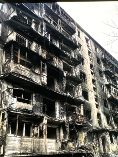 The image depicts a severely damaged apartment building in Neubrandenburg, Germany. The building appears to have sustained significant fire damage, evidenced by blackened facades, scorched balconies, and broken windows. Debris is visible on balconies and at the base of the structure. The exterior walls show signs of peeling paint and water damage, suggesting a substantial incident. The scene is desolate, with no people or active signs of life. The sky is overcast and a bare tree branch is visible in the upper right corner, indicating a cold season. The overall impression is one of destruction and abandonment.