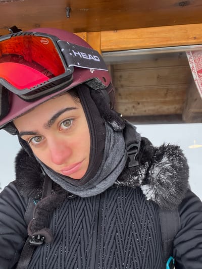 A close-up, eye-level image depicts an individual in Valtournenche, Italy. The subject wears a maroon 'HEAD' branded ski helmet with red mirrored 'RETHINK' branded goggles pushed up onto the helmet. A black balaclava is worn beneath a grey neck gaiter, covering the lower face, neck, and ears. The individual is dressed in a black winter jacket featuring a textured, chevron-quilted pattern on the body and a fur-lined hood or collar, which is lightly dusted with snow. A black backpack strap with a plastic buckle is visible over the individual's right shoulder. The subject has light-colored eyes and a complexion showing redness on the nose and cheeks, consistent with cold weather exposure. The background reveals a wooden building structure with horizontal planks, a visible metal fixture near the top, and a window reflecting a faint red and white patterned interior element. A bright, snowy environment is discernible behind the subject's left shoulder.