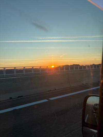 The image captures a view from a vehicle traveling on a highway during sunset. The sun, a bright orange orb, is low on the horizon, casting a warm glow across the sky. Several contrails are visible, streaking across the clear blue sky. A concrete barrier with a metal railing lines the side of the highway, separating the road from the landscape beyond. The landscape appears to be a flat expanse with a line of trees or hills in the distance, and a few windmills are visible. The road itself is dark asphalt with white lane markings. In the foreground, a side-view mirror of the vehicle reflects other vehicles on the highway. The overall impression is one of a journey during a picturesque sunset, likely on a major road. Based on the specific request for the location, this scene is situated on the A6 Autobahn near Braunsbach, Germany, a common route for travel through the region.