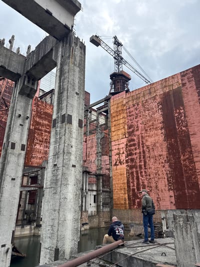 The image depicts an exterior view of Reactor 4 at the Chernobyl Nuclear Power Plant in Pryp'yat', Ukraine. Two people are visible on a concrete structure in the foreground. One person is standing and wearing a dark green jacket, blue jeans, and a baseball cap. The other person is leaning over the edge and wearing a black shirt and beige pants. In the background, the damaged reactor is visible, with its distinctive metallic red cladding and concrete support structure. A large crane is positioned above the reactor building. The sky is overcast.