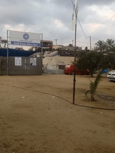 The image depicts a scene in Al-Mawasi, Palestinian Territories, under an overcast sky. The foreground consists of bare, light brown sand with scattered debris and a black hose or cable running diagonally from the lower left towards the lower right, passing a dark metal pole in the mid-right. A small, young palm-like plant is visible to the right of this pole.
In the midground, slightly to the left of the center, a large white rectangular sign with a blue border is prominently displayed on a metal frame. The sign features the "Médecins du Monde" (Doctors of the World) logo and the text "Médecins du Monde - France" in Latin script, with "أطباء العالم - فرنسا" (Doctors of the World - France) and "مركز رعاية صحية أولية" (Primary Health Care Center) in Arabic script below it. Below this sign, a dark grey corrugated metal gate or barrier with two white posters attached is visible.
Behind the sign and gate, numerous temporary structures and tents are visible. One prominent light-colored canvas tent clearly displays the words "FIRST AID" on its side. To the right of these tents, a red hatchback car is partially obscured. Further to the right, a white car with a visible license plate "D-964-G" is parked. Several green bushes and small trees are scattered around this area.
A tall, dark metal flagpole stands in the right midground, from which a white flag, partially obscured, flies. In the background, beyond the temporary structures, more permanent but worn buildings are visible on elevated ground, featuring rooftop water tanks. Palm trees are present on both the far left and far right sides of the background. No individuals are visible in the image.