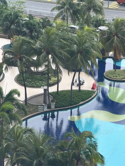 An aerial view captures a swimming pool area surrounded by palm trees and paved walkways. The swimming pool features a colorful mosaic design with sections of deep blue, light blue, and green. The water is slightly rippled, suggesting recent rainfall or movement. A red pool ladder with silver handrails is visible on the edge of the pool, next to a red safety barrier. Several small, floating white markers are spaced out in one section of the pool.
Surrounding the pool are lush green plants and manicured hedges. Paved pathways, with a light-colored tile pattern, curve around garden beds and lead to different areas of the pool. Several tall palm trees with broad fronds dominate the foreground and middle ground, their leaves creating dappled shadows. In one section of the paved area, a circular table is set with four small stools.
In the background, a road is visible with a yellow and white striped pedestrian crossing and a black metal fence. There are some vehicles on the road, including a dark-colored car and the back of a red truck. The overall environment suggests a tropical resort or hotel setting within the urban landscape of Subang Jaya City Council, Malaysia. The lighting indicates it is daytime, possibly overcast or after a rain shower. There are no visible people in the image.