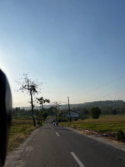 The image depicts a view from a motorcycle looking down a paved road in Nganjungganjung, Indonesia. The road curves slightly to the right and is flanked by green fields and sparse trees. In the distance, rolling hills covered in vegetation are visible under a clear blue sky. A few motorcycles are seen on the road ahead, with people riding them. The sun appears to be low in the sky, casting long shadows and creating a bright, hazy atmosphere. The foreground shows a portion of a dark object, likely the motorcycle's handlebar or helmet, on the left side of the frame. A single utility pole with wires runs parallel to the road.