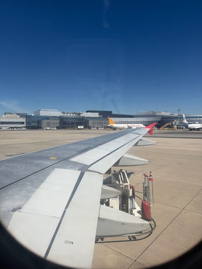 The image, taken from an airplane window, shows a view of Vienna International Airport in Schwechat, Austria. The foreground is dominated by the wing of the airplane the photo is taken from, which is white with grey accents and appears to be on the tarmac. In the middle ground, the airport terminal building is visible, with "FLUGHAFEN WIEN" and "VIENNA AIRPORT" signage clearly displayed. Several other aircraft are parked on the tarmac, including a white plane with "flypgs.com" branding and another with a blue and white livery. Various airport vehicles and equipment, such as a baggage tug and refueling equipment, are also present. The ground is a concrete tarmac, and the sky above is a clear, bright blue.