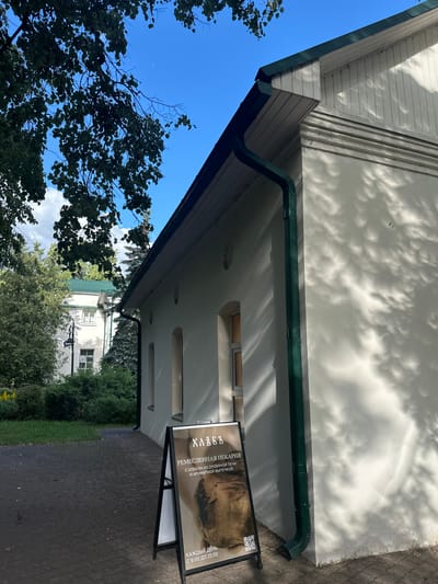 This is a photograph of a building with a green gutter and a white facade, dappled with shadows from trees. In the foreground, a black A-frame sign stands on a paved area. The sign displays Russian text and an image of bread. The text on the sign reads: "ХЛЕБЪ", "РЕМЕСЛЕННАЯ ПЕКАРНЯ", "С ХЛЕБОМ ИЗ ДРОВЯНОЙ ПЕЧИ И АРОМАТНОЙ ВЫПЕЧКОЙ", and "КАЖДЫЙ ДЕНЬ С 9:00 ДО 21:00". This translates to "BREAD", "ARTISAN BAKERY", "WITH BREAD FROM A WOOD-BURNING OVEN AND FRAGRANT PASTRIES", and "EVERY DAY FROM 9:00 TO 21:00". The sign indicates that this is an artisan bakery. Beyond the building, there are trees and another building visible. The sky is blue and clear, suggesting it is daytime with good weather. The scene appears to be outdoors, possibly in a town or city in Russia, given the location context. The overall impression is of a bright, sunny day outside a local establishment.