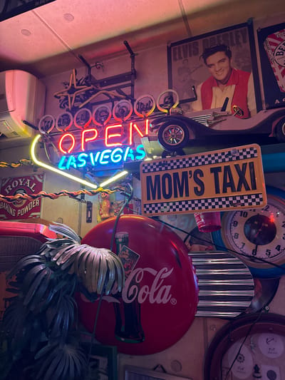 This media file shows a cluttered display of memorabilia, likely in a themed establishment. The scene is dimly lit, with a strong focus on neon signs and vintage items.
Dominant subjects include a neon sign that reads "OPEN LAS VEGAS" in alternating red and blue letters, with yellow accents. Another neon sign, shaped like a license plate, displays "MOM'S TAXI" in yellow and black. A framed picture of Elvis Presley, with him wearing a red jacket, is prominently featured. A large red Coca-Cola sign is visible, along with a vintage clock. Other objects include decorative tin items, a toy car wheel, and what appear to be artificial plants. There's also a red can with "CU" visible, and a box labeled "ROYAL BAKING POWDER." The background appears to be a textured wall.
No clear activity or event is taking place; it's a static display. The overall atmosphere is retro and potentially a bit kitschy, evoking a nostalgic or entertainment-focused environment. The lighting suggests it could be nighttime or an indoor setting with artificial light. There are no explicit weather cues. The text "LAS VEGAS" on the neon sign provides a location cue, although the provided location context is Adachi, Japan, suggesting the display might be themed. The emotion conveyed is likely one of fun and nostalgia.