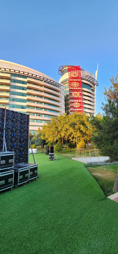 The image depicts an outdoor setting in front of a modern, curved, white building with numerous windows. A large, red and gold tapestry or banner with intricate patterns is prominently displayed on the building's facade, likely for a cultural event or celebration. In the foreground, a large LED screen or video wall is set up on the vibrant green artificial turf, along with several black speaker cases, suggesting preparations for an outdoor event such as a concert or festival. Trees with lush green foliage and some with golden-yellow leaves are visible behind the screen, indicating it might be autumn or late summer. The sky is a clear, bright blue, suggesting a sunny day. The overall scene captures a blend of modern architecture and outdoor event setup, with a touch of cultural decoration. The orientation of the image is rotated.