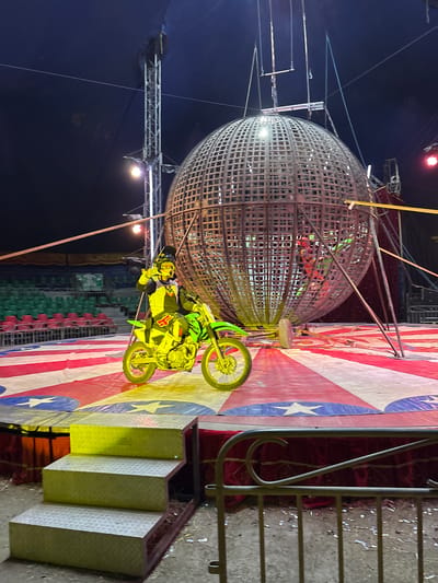 The image shows a circus act in progress. A motorcyclist dressed in yellow riding gear is on a yellow motorcycle. They are on a stage decorated with red and white stripes and blue stars reminiscent of the American flag. A large spherical cage, known as the "Globe of Death," is present, with another motorcyclist visible inside. The location is identified as Tejalpa, Mexico. In the background, there are stadium-style seats, some empty, in shades of green and red. A metal scaffolding structure and various spotlights are also visible, suggesting an outdoor circus venue. A set of metal stairs leads up to the stage.