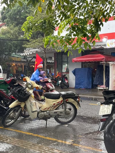 This image captures a street scene in Ngũ Hành Sơn Ward, Vietnam, during a light rain shower. The asphalt pavement is wet, exhibiting reflections and partially visible yellow road markings.
In the foreground, a cream-colored, possibly Daelim, motorbike with a black seat is parked on its kickstand, facing left. It has a black wire basket on the front handlebars holding dark items and a transparent plastic bag hanging from the front. To its right, the rear section of a dark motorbike is visible, displaying a license plate "37-B2 790.63".
A person wearing a blue rain poncho and an orange helmet is actively riding a dark-colored motorbike from left to right, accompanied by a passenger in a purple rain poncho. Their forms are slightly blurred due to motion.
Along the right side of the street, a row of commercial buildings is present. One storefront shows a sign for "NTLEY coffee." Another building features a white facade with red trim. A red Vietnamese flag with a yellow star is prominently displayed on a pole. Further right, a red awning extends over a sidewalk area, beneath which a blue tarp covers an unseen object or stall. Green leafy tree branches overhang the scene from the top, with visible raindrops evident throughout the image.