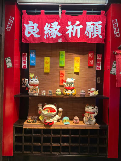 The image displays a retail or display alcove, framed by prominent red panels, characteristic of a commercial establishment in New Taipei City, Taiwan. A woven bamboo mat serves as the central background for two dark wooden display shelves. Above, a red fabric banner with white Japanese characters reads "良緣祈願" (Prayer for good match/relationship). Six colored vertical strips are suspended against the bamboo, each bearing a single Japanese word: blue for "健康" (Health), yellow for "厄除" (Ward off evil), green for "合格" (Passing an exam/success), orange for "恋愛" (Love), another yellow for "富貴" (Wealth/Prosperity), and red for "幸福" (Happiness).
The top shelf presents a collection of figures: a white Maneki-Neko (beckoning cat) with a raised left paw and a price tag of "1500," a larger, stacked figure resembling a Maneki-Neko atop a Daruma doll, inscribed with "大勝" (Big Win), two miniature Maneki-Neko enclosed in clear boxes, a green Daruma doll, and two additional small, egg-shaped figures.
The bottom shelf holds a similar assortment: a large white Maneki-Neko with both paws raised, two small items in clear cases, a blue cap adorned with a sunflower design, a pink egg-shaped figure, and another white Maneki-Neko with a raised right paw and a price tag of "800."
Affixed to the red side panels are several white-on-red stickers reading "良緣達成" (Good match achieved) and a green sticker on the left panel marked "大豐" (Great harvest/abundance). The lower portion of the display features a dark wooden cubby shelving unit with multiple square compartments. No people are visible; the scene is a static arrangement of merchandise.