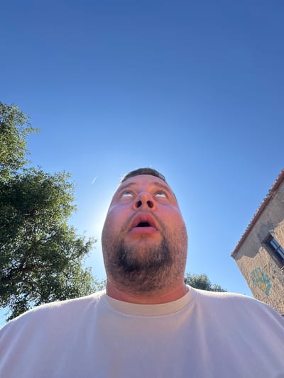 The image is a low-angle, wide shot of a man in Santa Cristina d'Aro, Spain. The man, who appears to be an adult male with facial hair, is wearing a white t-shirt and looking up at the camera with an astonished expression. His mouth is open, and his eyes are wide. The sun is bright, suggesting it is daytime, and the sky is a clear blue with a contrail from an airplane visible.

In the background, a building with a tiled roof and a window can be seen at the top of the frame. The building has some graffiti on its exterior wall. To the right of the building, the leaves of a tree are visible, also illuminated by the sunlight. The overall setting appears to be outdoors, possibly in a rural or suburban area, given the presence of the building and trees against a clear sky.

The man's expression and the angle of the photo suggest a playful or surprised moment. The bright sunlight and clear sky indicate fair weather. No other individuals are visible in the frame. There is some blue and yellow spray paint graffiti on the building, but no other discernible text.