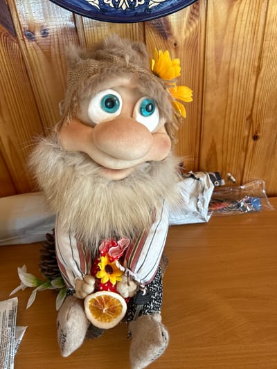 The image shows a close-up of a handmade doll sitting on a wooden surface. The doll is designed to resemble a gnome or a similar mythical creature, with a large nose, prominent eyebrows, and a fluffy beard. It has bright blue eyes and is wearing a striped shirt, patterned pants, and a woven hat adorned with a yellow flower. The doll is holding a small bunch of items, including a sunflower, a red polka-dotted fabric, and a slice of dried orange. To the left of the doll, there are some white flowers and a small white packet with text in Russian. Behind the doll, there is a stack of white paper packaging, and beyond that, a wooden wall. The overall setting appears to be indoors, possibly on a shelf or table. The location is not specified within the image.