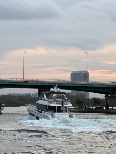 A white motor yacht, named "NENA," cruises through choppy water under a cloudy sky. The yacht is moving away from the camera, creating a significant wake. A large bridge, painted green, spans across the frame in the background, with lampposts visible on either side. Beyond the bridge, a modern, light-colored building with many windows stands on a tree-lined shore. The overall scene suggests a daytime activity in a coastal urban area, likely Bal Harbour Village, Florida, given the presence of a luxury yacht, a bridge, and the distinctive architecture.