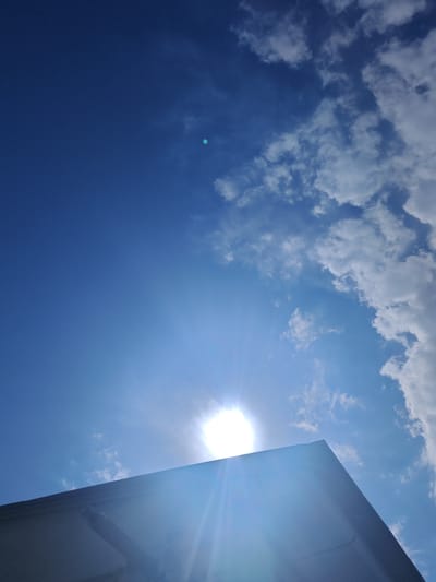 The image captures a bright, sunny day with a clear blue sky and scattered white clouds. The sun is prominently visible, casting a strong glare and lens flare across the scene. In the foreground, the sharp edge of a dark, angled structure, possibly a roof or building, cuts across the lower portion of the frame. This structure is silhouetted against the bright sky. The sun's rays extend outward, creating a starburst effect. The overall mood is one of a clear, possibly hot, day, with the sun dominating the visual field. The location is identified as Jaukаshty, Uzbekistan. There is no visible text in the image.