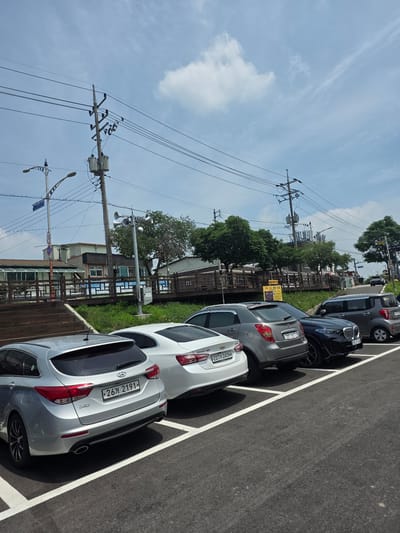 This is a daylight, eye-level, outdoor, medium shot of a parking lot in Anseong-si, South Korea. Six cars are parked in a row on an asphalt surface marked with white parking lines. The cars are, from left to right: a silver Hyundai i40 wagon, a white Chevrolet Malibu, a gray SsangYong Tivoli, a black BMW X6, a gray Kia Morning, and a white Hyundai Avante. In the background, there is a wooden deck with stairs leading up to a building with a green roof and glass windows. Beyond the building, trees and utility poles with power lines are visible against a bright blue sky with scattered clouds. A street sign is visible on a pole to the left of the wooden deck. The overall scene depicts a typical urban parking area with surrounding infrastructure under a daytime sky.