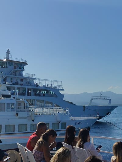 The image shows two ferries docked in the port of Glyfa, Greece. The foreground ferry, named "VENUS", is partially visible, with several passengers seated on deck. The passengers are diverse in age and gender, and some are interacting with each other or looking at their phones. The ferry is white with blue trim, and it has multiple decks and windows. Behind the "VENUS" ferry, a larger, grey ferry is docked. This ferry has a more industrial appearance, with cranes and other equipment visible on its deck. The background shows a clear blue sky and distant, hazy mountains. The water is calm and reflects the sunlight. The overall scene is one of a busy port on a sunny day, with ferries arriving and departing and passengers waiting to board or disembark.