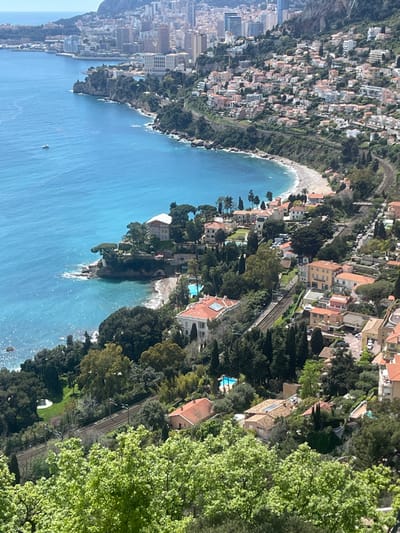 The image is an aerial view of Roquebrune-Cap-Martin, France, taken from a hillside overlooking the Mediterranean Sea. The image is focused on the coastline, with the foreground dominated by lush greenery and a few houses. In the background, a city sprawls up the hillside, with buildings of varying sizes and a prominent rocky outcropping. The coastline is characterized by a sandy beach, turquoise water, and rocky inlets. A small boat is visible on the water. There is a train track snaking its way along the hillside, with cars and other vehicles parked along its side. The image is taken from the viewpoint with the coordinates 43.763 latitude and 7.464 longitude. The image was likely taken on a sunny day, with clear skies and a bright blue sky. The composition is balanced, with the foreground providing a sense of depth and the background providing context for the location. There are no people visible in the image.
