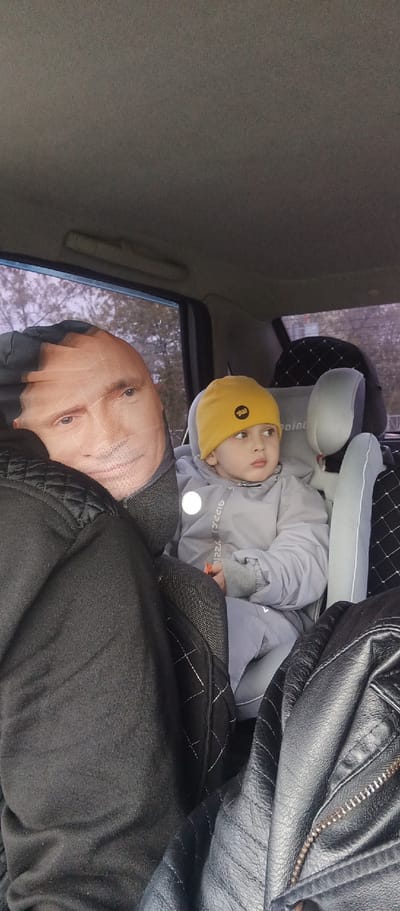 Inside a vehicle, a young child with brown hair and eyes, wearing a yellow knit hat and grey snowsuit, is seated in a grey child car seat. The child is looking to the right. To the child's left is an adult whose face is obscured by a pillow printed with the face of Vladimir Putin. Various jackets and bags are also visible in the vehicle. The environment visible through the window suggests an urban setting in Irbit, Russia.