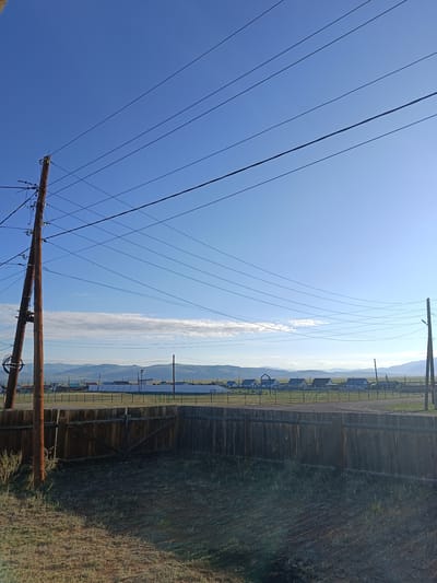 The image depicts a rural scene under a clear blue sky with a few wispy clouds in the distance. A cluster of houses, some with red roofs, are visible behind a dark wooden fence in what is identified as Zhana-Aul, Russia. The fence runs horizontally across the foreground, with a gate visible on the left. Several power lines stretch across the sky, supported by wooden utility poles, one of which is prominently in the left foreground. The landscape beyond the houses appears to be rolling hills or mountains. The overall impression is one of a quiet, possibly remote, residential area.
