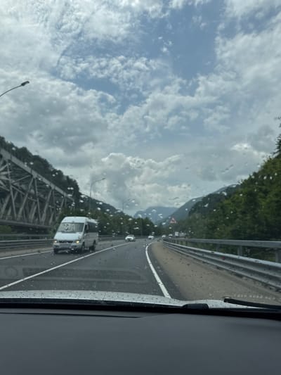 The image depicts a view from inside a vehicle driving on a highway, possibly in or near Chvizhepse, Russia, given the context. The windshield is covered in raindrops, indicating overcast or rainy weather. The highway has multiple lanes and is flanked by a metal guardrail on the right. On the left side of the highway, there is a large, grey metallic bridge structure, likely a railway bridge, spanning across a mountainous terrain covered in lush green trees. In the distance, mountains are visible under a cloudy sky with patches of blue. Several vehicles are on the highway, including a white minibus in the foreground and other cars further ahead. A streetlamp is visible on the left side of the road, and a traffic sign indicating a sharp turn is visible in the middle distance on the right side of the highway. The dashboard of the vehicle is visible at the bottom of the frame.