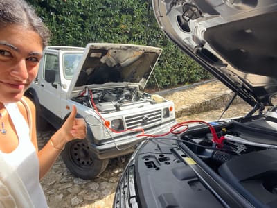 The scene takes place outdoors in Vila Nova de Cerveira, Portugal. A female subject with dark hair is partially visible on the left side of the frame, smiling and giving a thumbs-up with her right hand. She wears a light-colored sleeveless top and a thin bracelet on her right wrist, with a small charm visible on a necklace. Two vehicles are positioned on a paved surface composed of irregular stones.
On the left, a white Suzuki Samurai 4x4 is parked with its hood open, revealing its engine compartment. Red and black jumper cables are connected to terminals within this vehicle's engine bay. On the right, partially in the foreground, is a dark-colored modern SUV, also with its hood open to expose its engine bay. The red and black jumper cables extend from the white Suzuki Samurai to terminals within the dark SUV's engine compartment, indicating a battery jump-start in progress. Lush green foliage forms the background, suggesting a sunny outdoor environment.